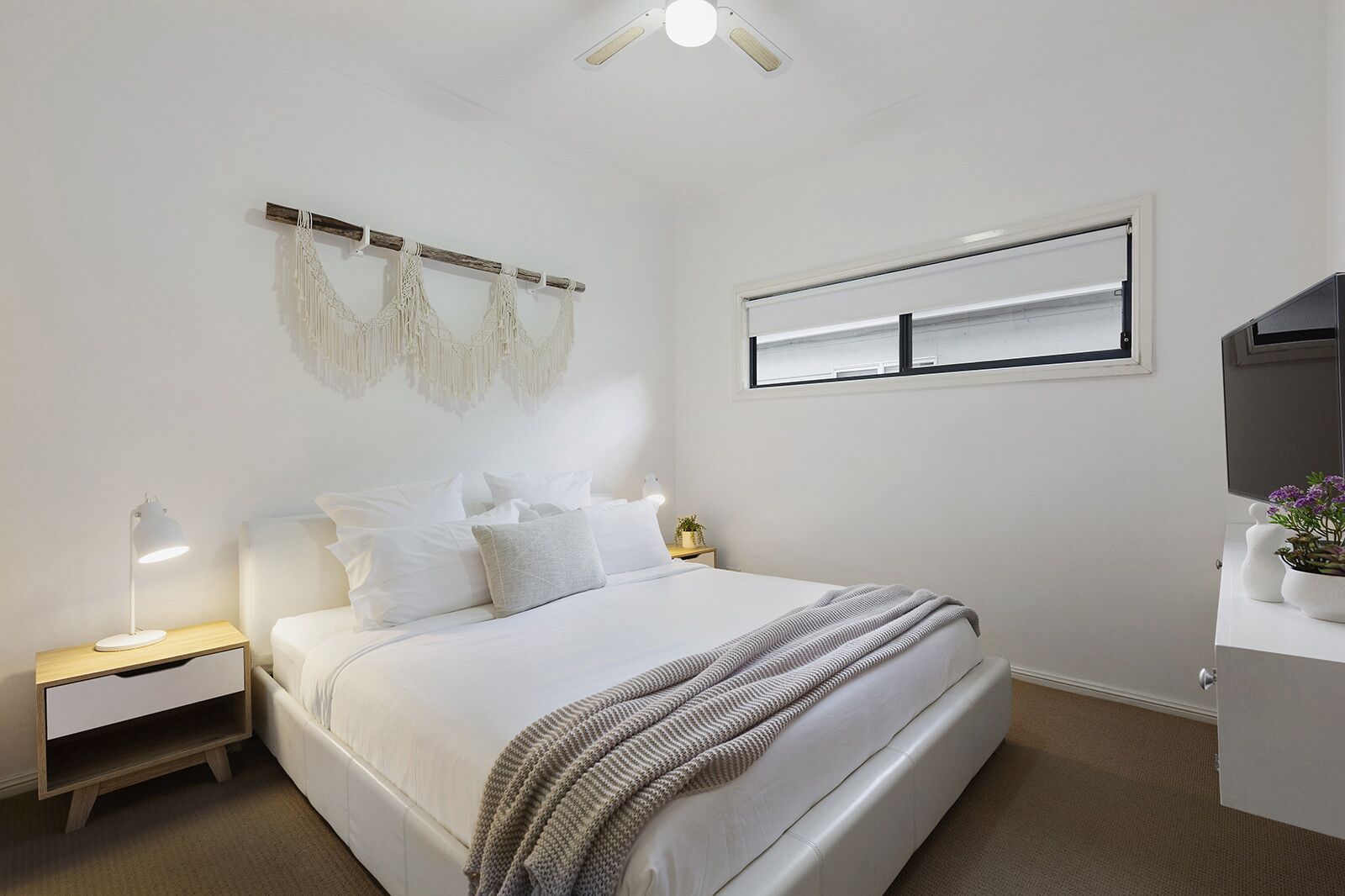 A modern bedroom with a large bed, a bedside table with a lamp, a wall-mounted macrame piece, a window, and a flat-screen TV on a cabinet.