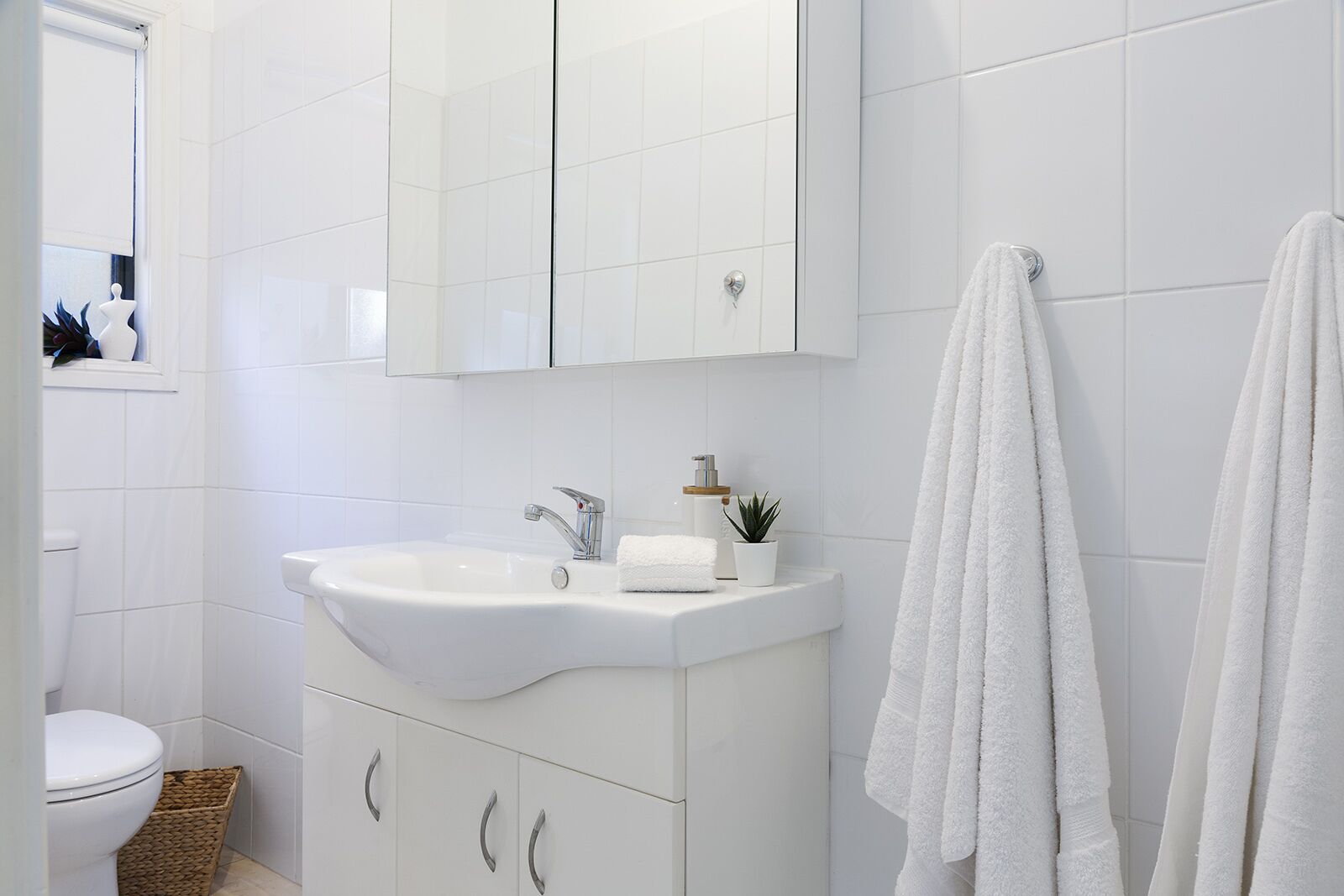 A modern bathroom with white tiles, a white sink, and a mirror above it. There is a towel hanging on the wall and a small plant on the sink counter.