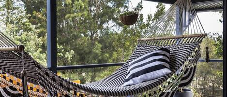 A hammock chair with a striped cushion, a hanging plant, and a view of trees through large windows.