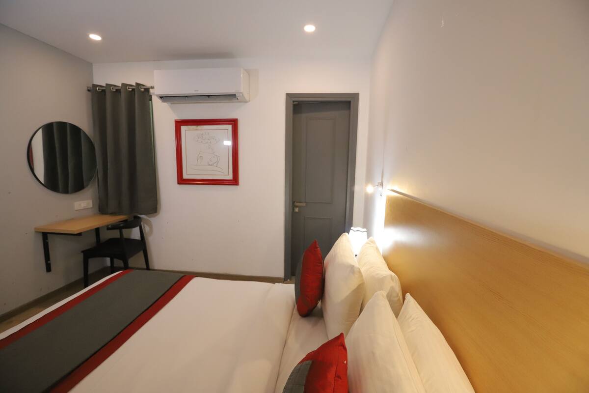 A hotel room with a bed, a desk, a chair, a round mirror, a framed picture, and an air conditioning unit.