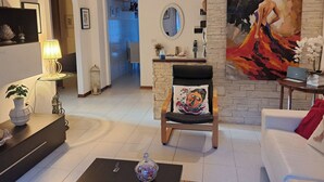Een moderne woonkamer met een bank, een fauteuil met een kleurrijk kussen, een salontafel, een lamp en een schilderij aan de muur.