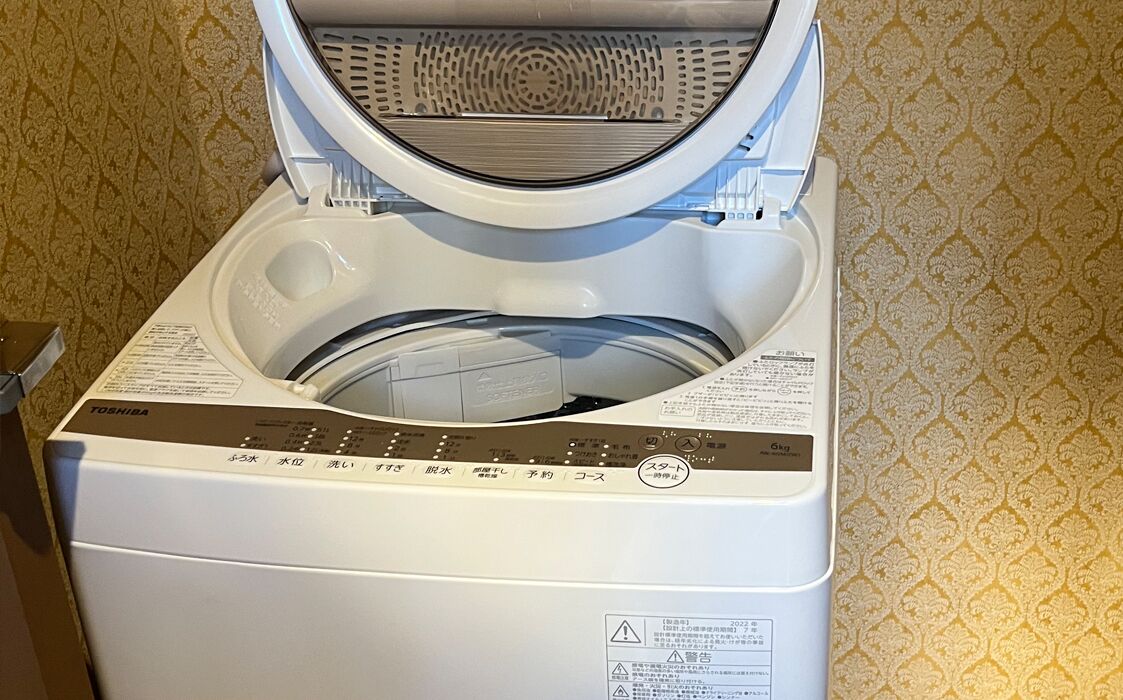 Vista frontal de una lavadora blanca con panel de control y una etiqueta en japonés.