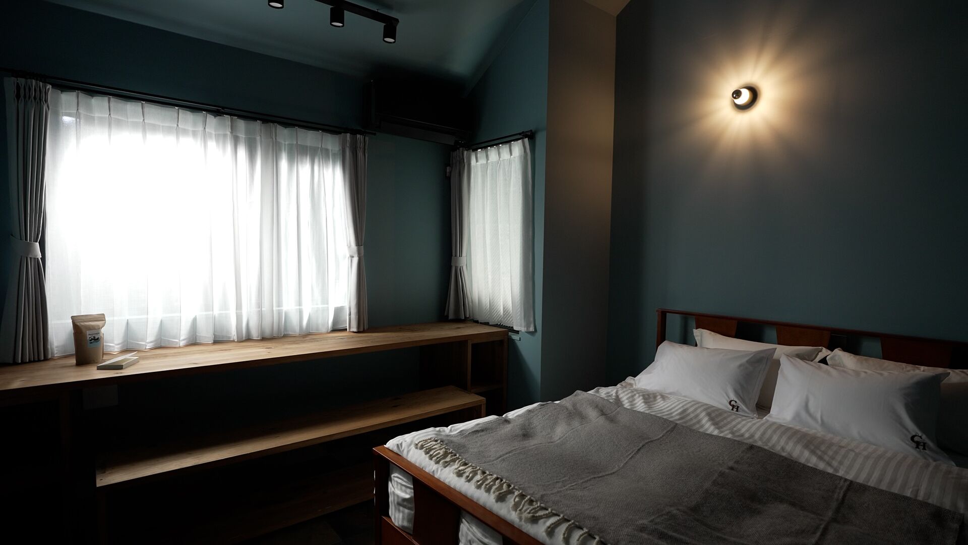 A bedroom with a bed, a bedside table, a window with curtains, and a wall-mounted light.