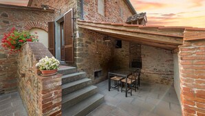 Un pati exterior rústic amb un mur de maó, unes escales de fusta, una taula amb cadires i plantes en testos.