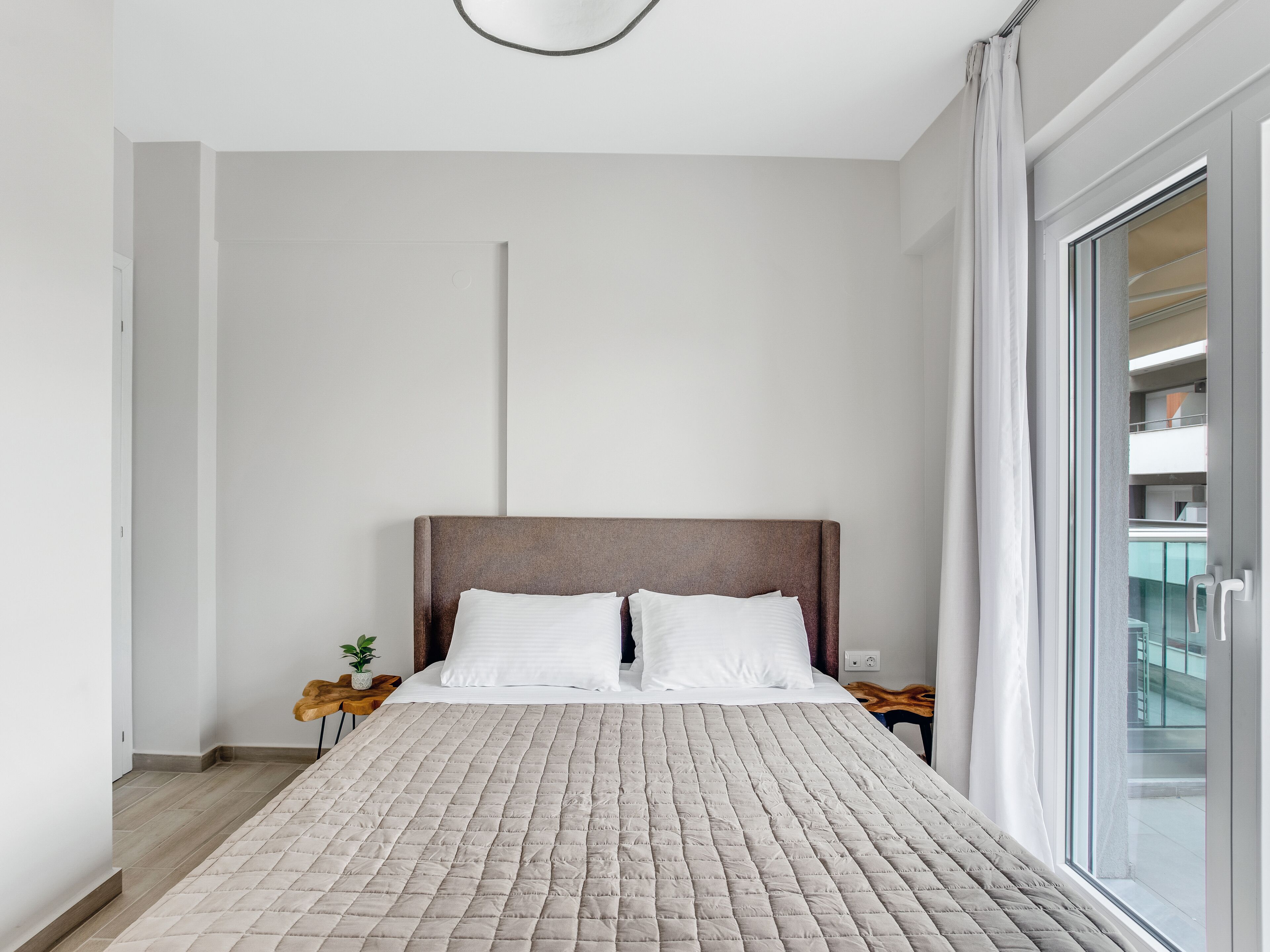 Un dormitorio moderno con una cama grande, una mesita de noche de madera y vistas a un edificio a través de un ventanal.