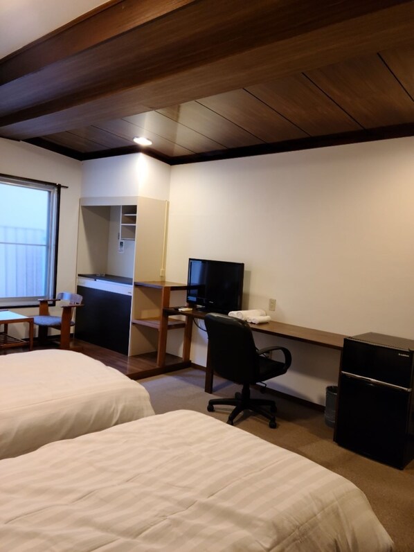 A hotel room with two beds, a desk with a computer, a chair, a small refrigerator, and a window.