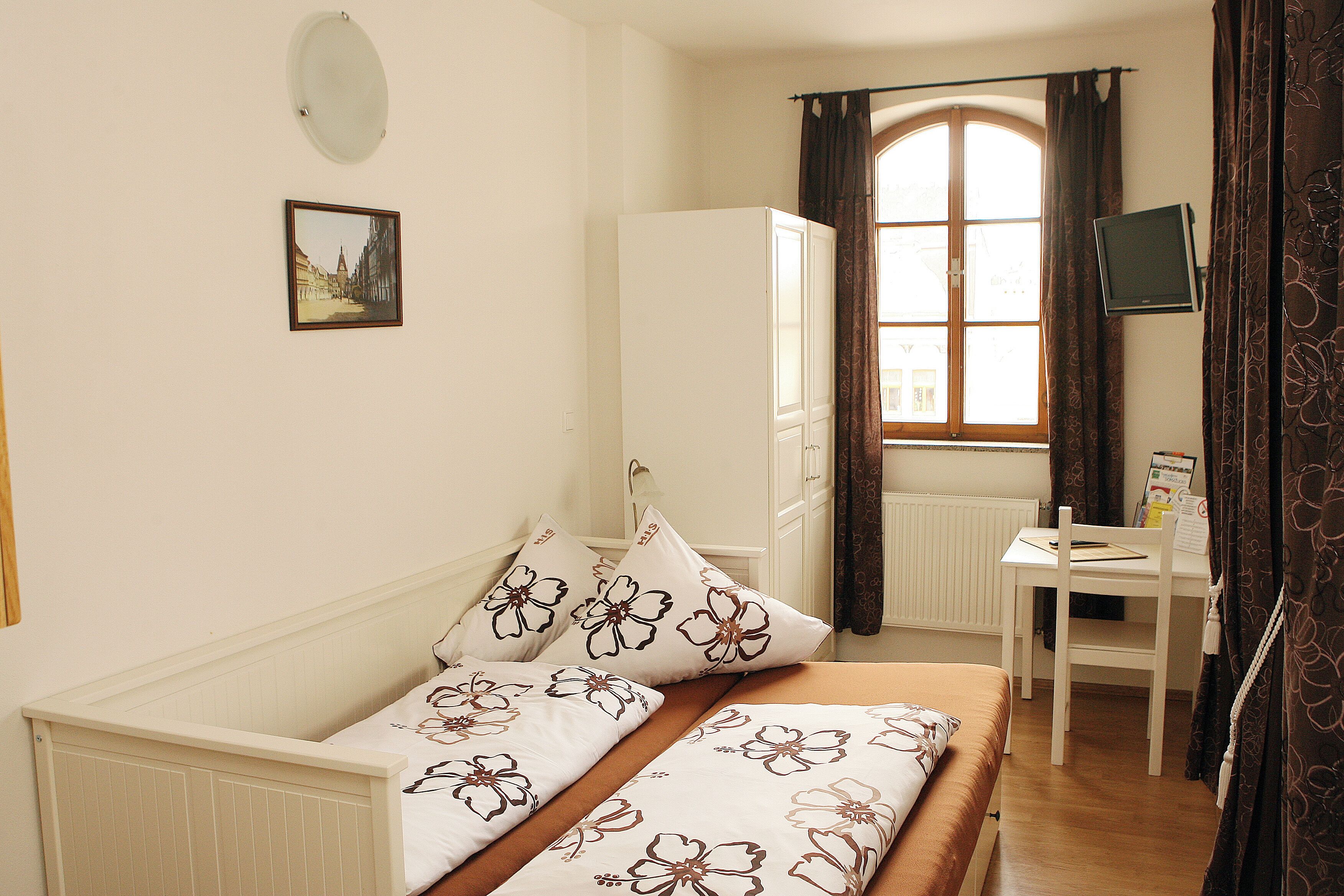 Ein Schlafzimmer mit Bett, Schreibtisch, Kleiderschrank, einem Fenster mit Vorhängen und einem Wandbild.