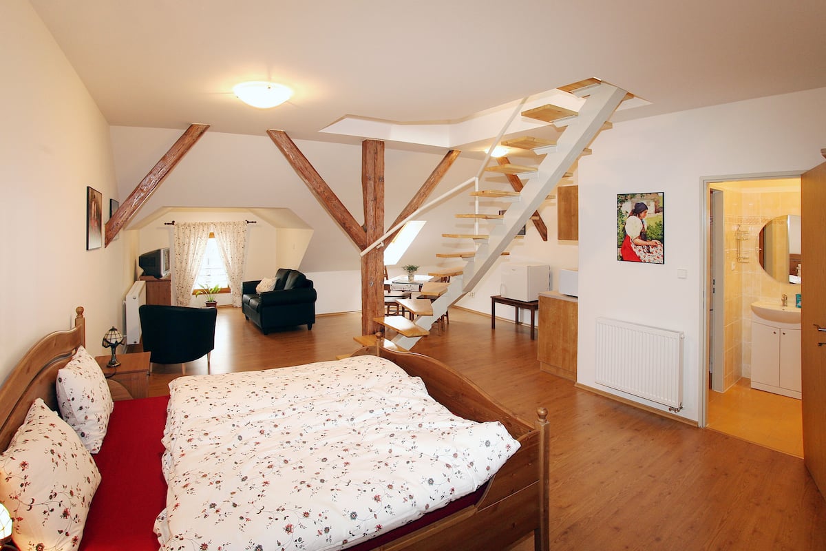 Een slaapkamer met een houten bed, een rood tapijt, een trap en een badkamer met een wastafel en spiegel.