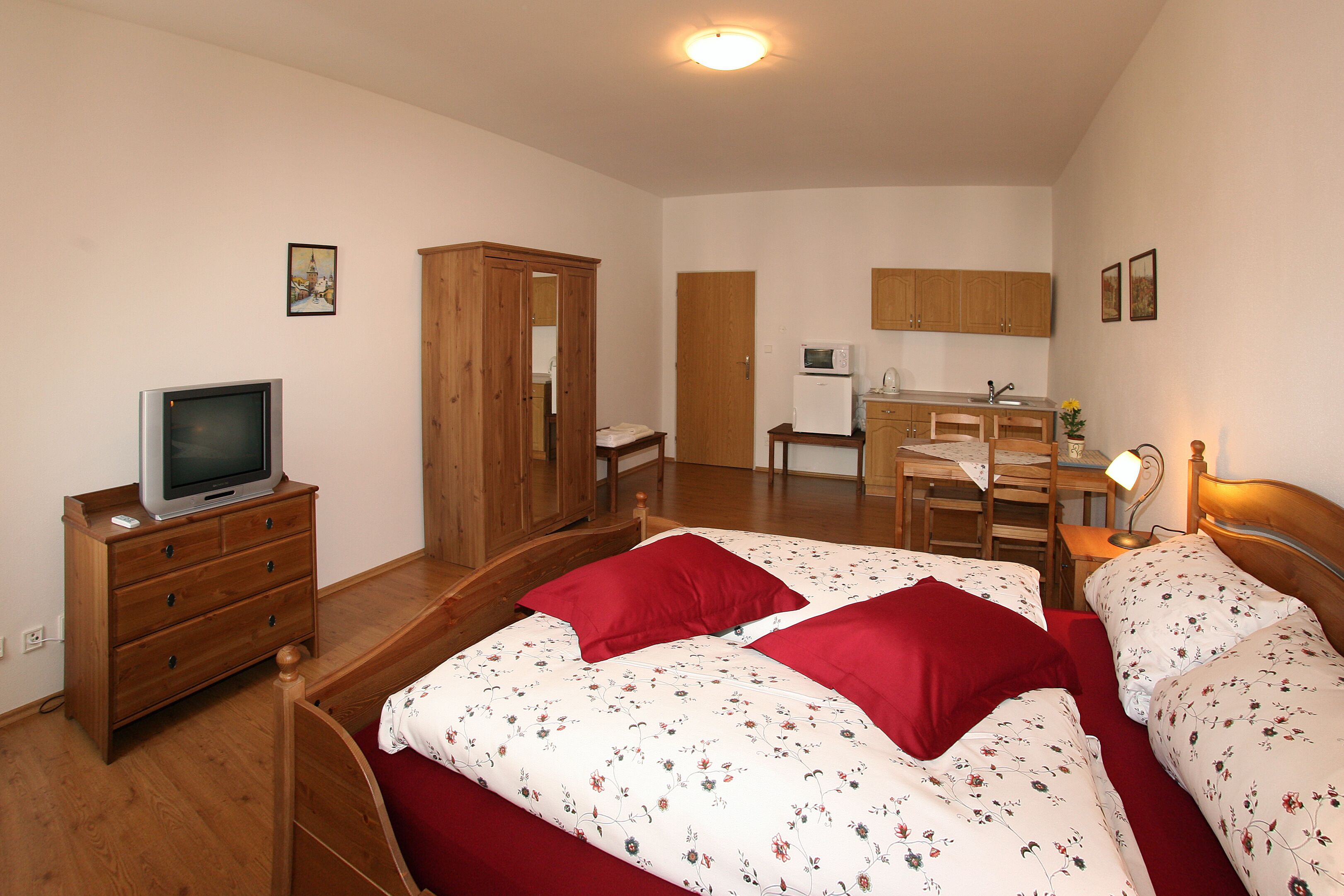 Un dormitorio con dos camas, un armario de madera, un televisor sobre una cómoda y una kitchenette al fondo.