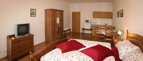 Un dormitorio con dos camas, un armario de madera, un televisor sobre una cómoda y una kitchenette al fondo.