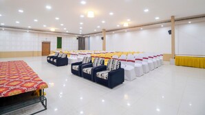 A spacious conference room with rows of white chairs, a long table with a patterned tablecloth, and a stage area with a yellow backdrop.