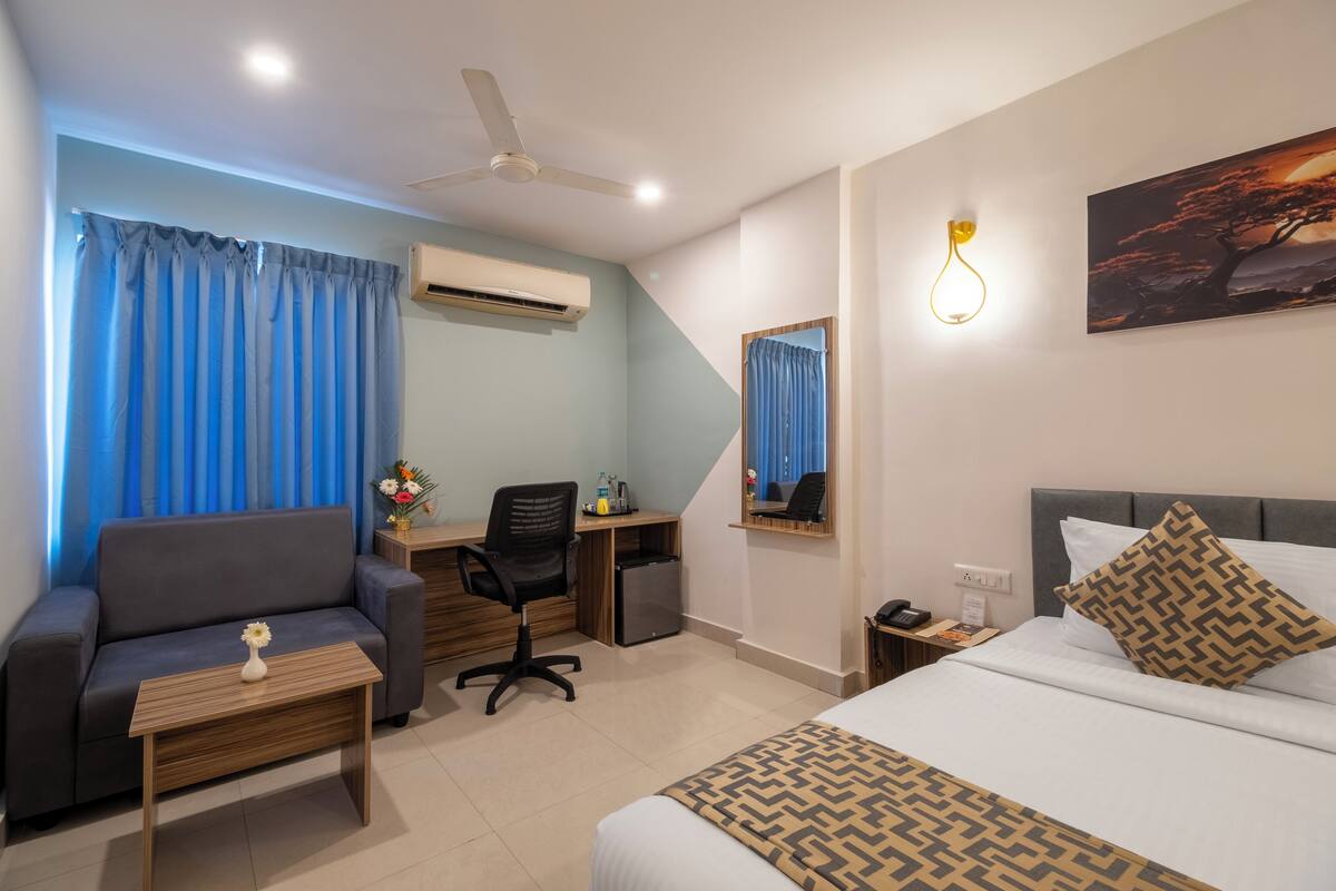 A hotel room with a bed, a sofa, a desk, a chair, a small table with a vase, a ceiling fan, an air conditioning unit, a nightstand, a lamp, and a painting on the wall.
