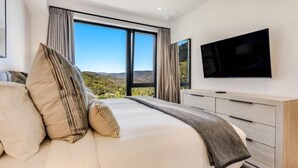 A modern bedroom with a large bed, a flat-screen TV, and a scenic view through the windows.