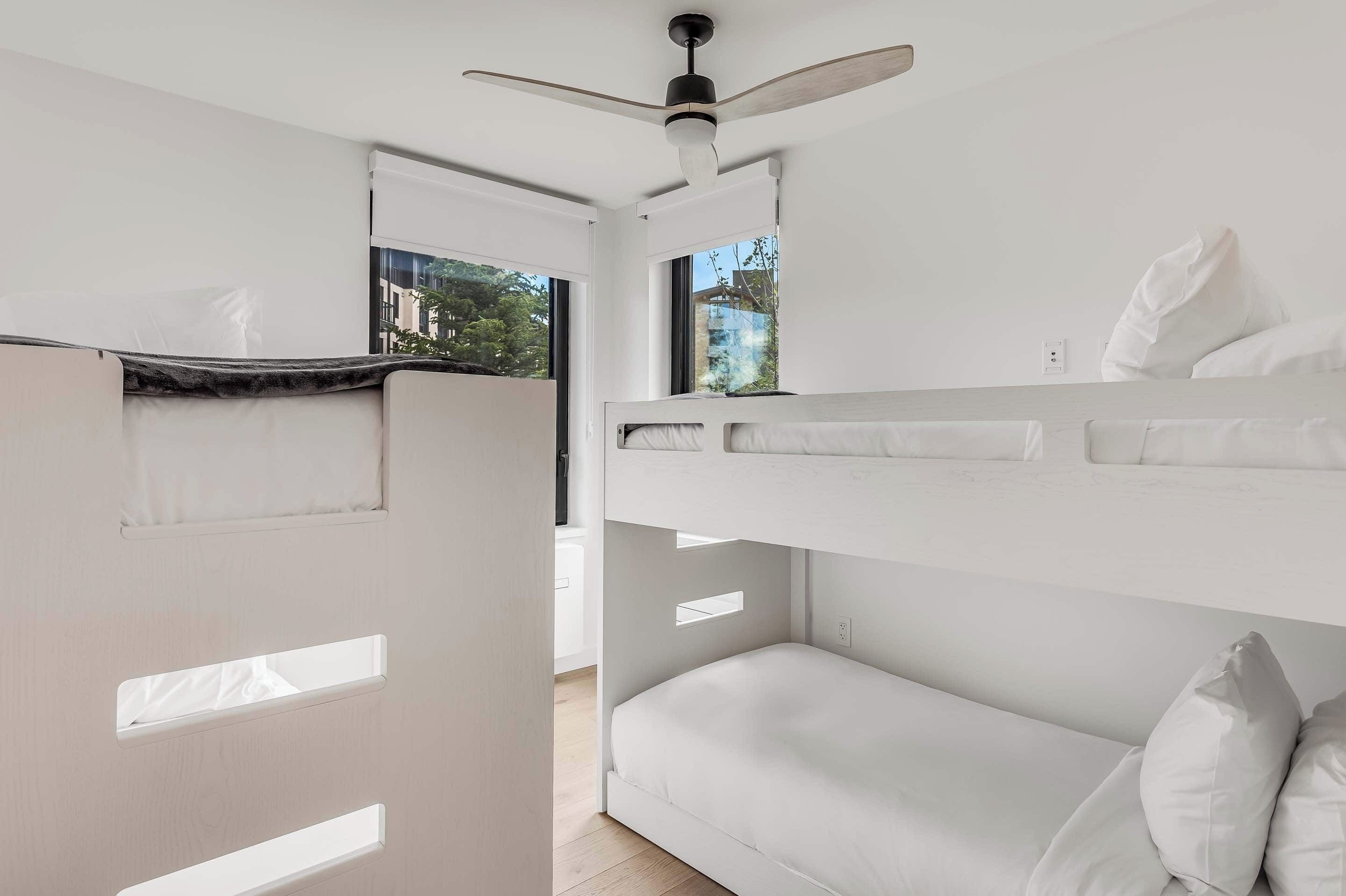 Une chambre moderne et minimaliste, dotée de lits superposés, d’un ventilateur de plafond et de grandes fenêtres offrant une vue sur l’extérieur.