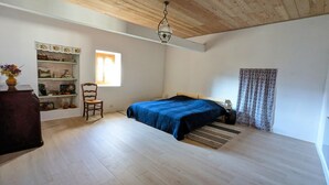 A bedroom with a wooden floor, a bed with a blue bedspread, a wooden chair, a shelf with decorative items, and a window with curtains. - Vacation rental lakes, vineyards and mountains area (MASSIGNIEU DE RIVES)