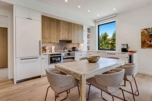A modern kitchen with white cabinetry, a wooden accent wall, a large window, and a dining area with a marble table and upholstered chairs.