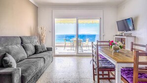 Un salón con un sofá gris, una mesa de comedor de madera con sillas, una televisión y vista al mar a través de un ventanal.