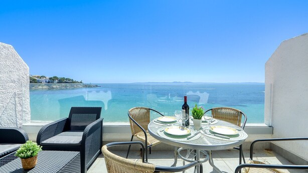 Un balcón con mobiliario de mimbre, una mesa dispuesta para comer y vista al mar.