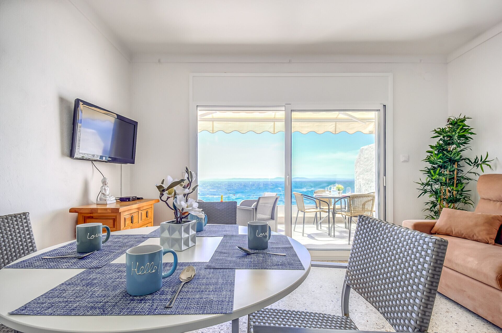 A dining area with a table set for two, a television mounted on the wall, and a view of the sea through large sliding doors.