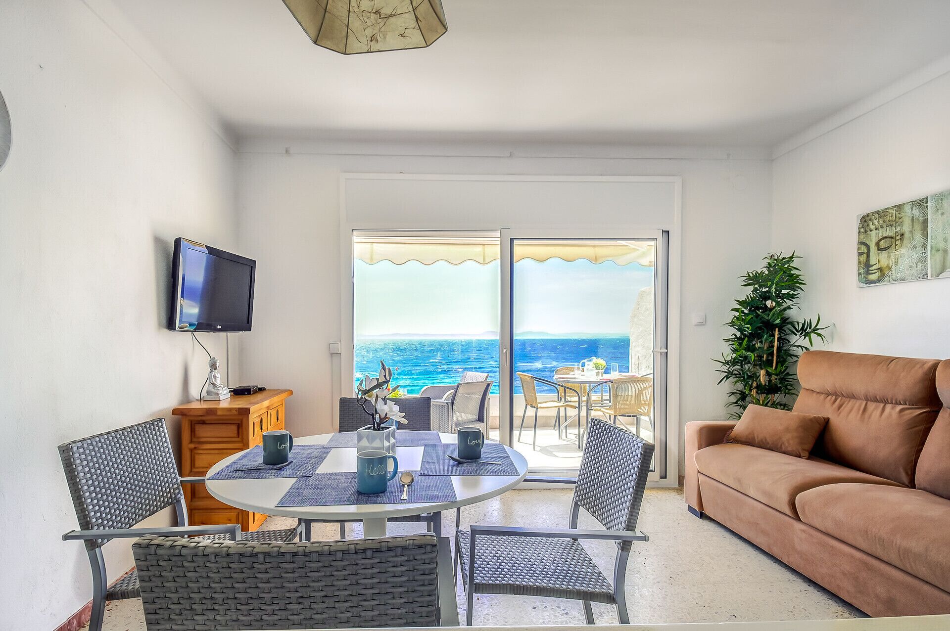A living room with a sofa, a TV, a dining table with chairs, and a view of the sea through a glass door.