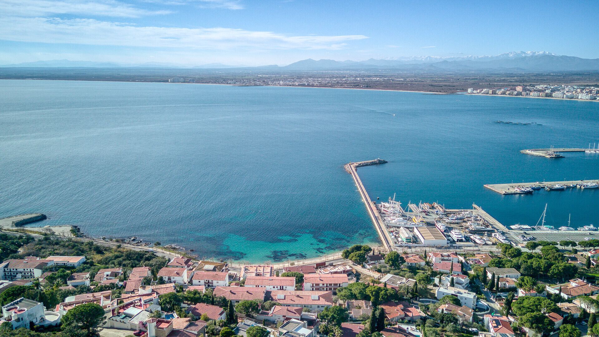 An aerial view of a coastal town with a marina, clear blue waters, and a mountainous backdrop.