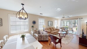 A spacious living room with a dining area, large windows with blinds, a chandelier, and a ceiling fan. - Golf Course Oasis Inviting 3BRVilla Pawleys Island (Pawleys Island)
