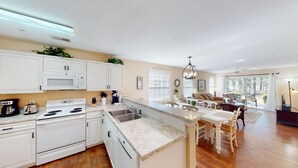 A spacious kitchen with white cabinetry, a built-in oven, and a large island with a sink. There is a dining area with a table and chairs, and a living room with a sofa and a large window. - Golf Course Oasis Inviting 3BRVilla Pawleys Island (Pawleys Island)