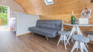A cozy room with a grey sofa, a small round table with a bottle and glasses, and a wicker basket. - High Farm Deluxe Pod (Pet) (Beverley)