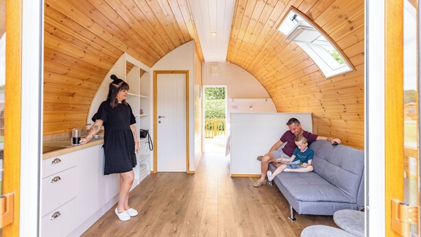 A cozy interior with wooden walls, a kitchenette, a sofa, and a door leading outside. - High Farm Deluxe Pod (Pet) (Beverley)