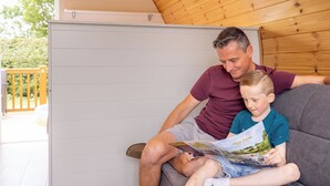 A man and a child sitting on a grey couch in a room with wooden paneling and a whiteboard. - High Farm Deluxe Pod (Pet) (Beverley)