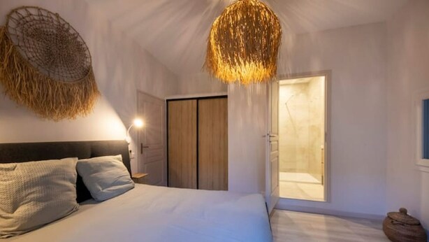 A bedroom with a bed, a nightstand, a wall-mounted lamp, a wooden wardrobe, and a door leading to a bathroom. - Sublime 2 Pièces Avec Jardin (Villefranche-sur-Mer)
