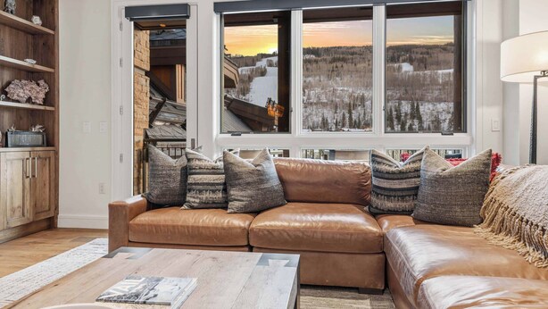 A cozy living room with a brown leather sofa, a wooden coffee table, and a large window with a view of a snowy landscape. - Solaris 6D East - 4 Bedroom + Den (Vail)