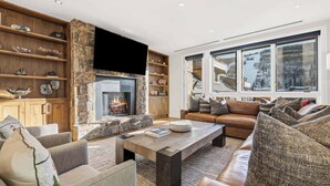 A cozy living room with a stone fireplace, leather sofas, and a large window offering a view of a snowy landscape. - Solaris 6D East - 4 Bedroom + Den (Vail)