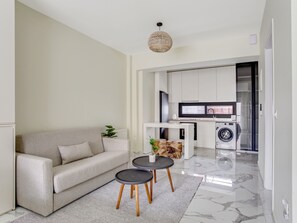 A modern living room with a sofa, two small tables, and a kitchen area with a washing machine. - Sanders Old Port - Simple Studio w/ Balcony (Thessaloniki)
