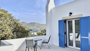 Una terraza con mesa y sillas, un edificio blanco con contraventanas azules, y vistas al mar y las montañas.