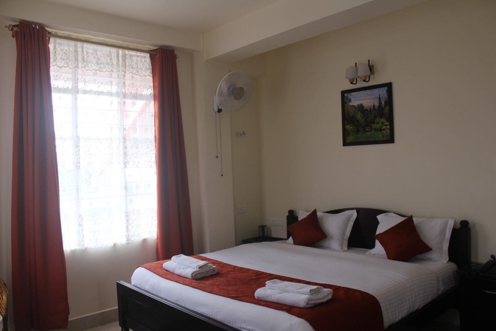 Una habitación de hotel con cama, ventilador de pared, un cuadro y ventana con cortinas.