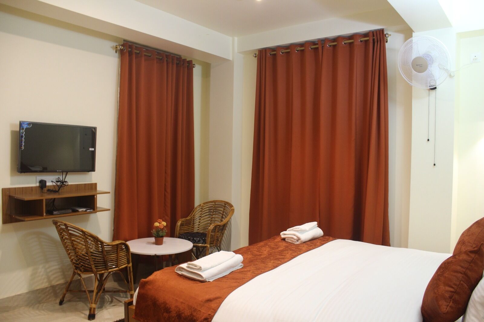 A hotel room with a bed, two chairs, a small table, a TV, and a wall fan.