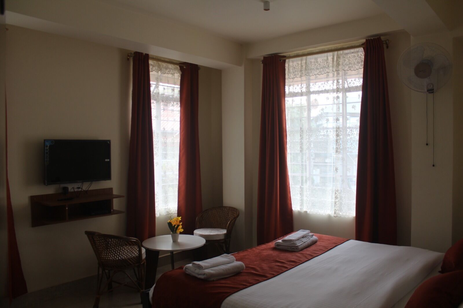 A hotel room with a bed, a TV, a small table with a vase of flowers, two chairs, and a window with curtains.