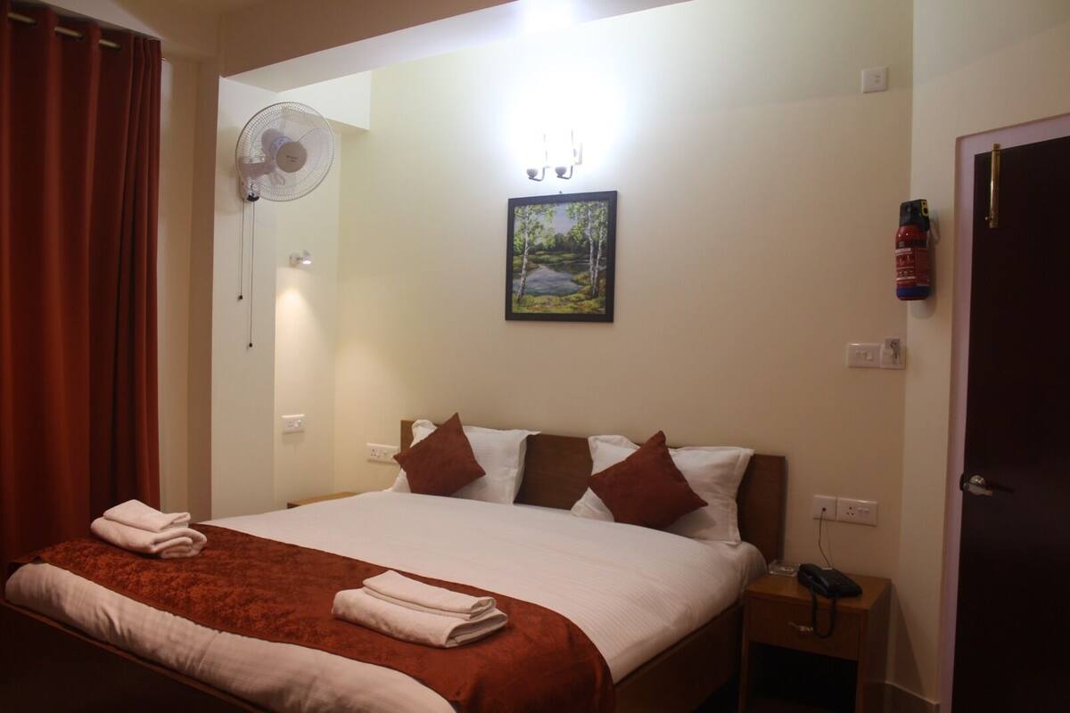 A hotel room with a bed, a bedside table with a phone, a wall-mounted fan, and a painting.