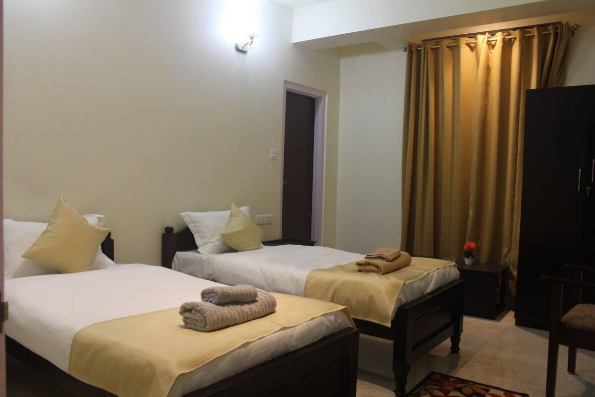 A hotel room with two beds, each with a beige bedspread and pillows, a small table with a plant, and a curtain on the window.