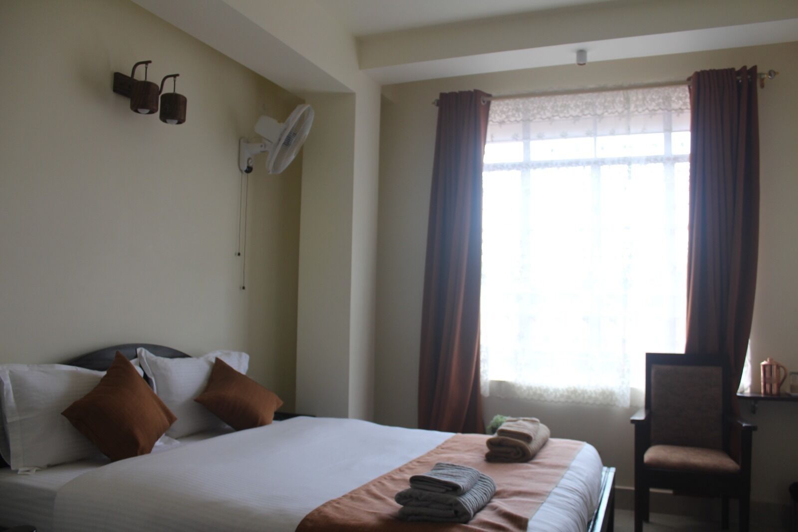 A hotel room with a bed, pillows, a desk, a chair, a window with curtains, and wall-mounted fans.
