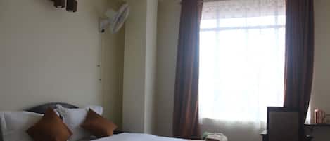 A hotel room with a bed, pillows, a desk, a chair, a window with curtains, and wall-mounted fans.