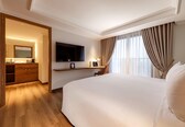 A hotel room with a large bed, a TV mounted on the wall, a desk with a lamp, and a wooden wardrobe.