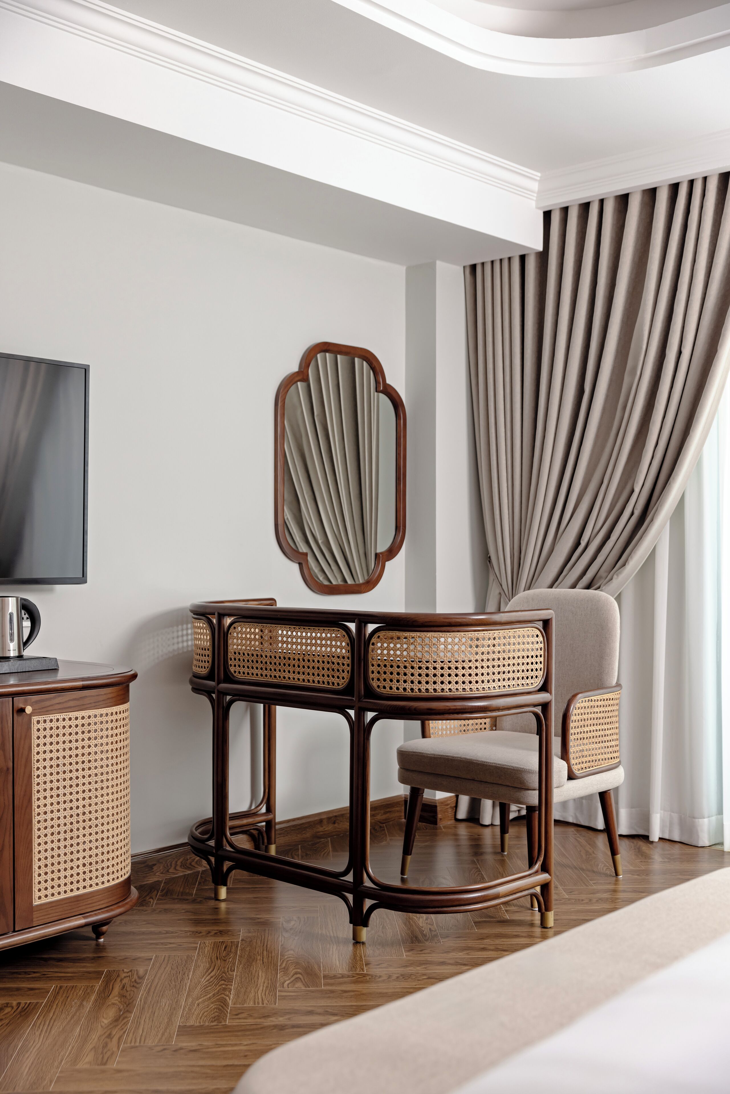 A hotel room with a wooden floor, a television, a rattan side table, a chair, and a large window with curtains.