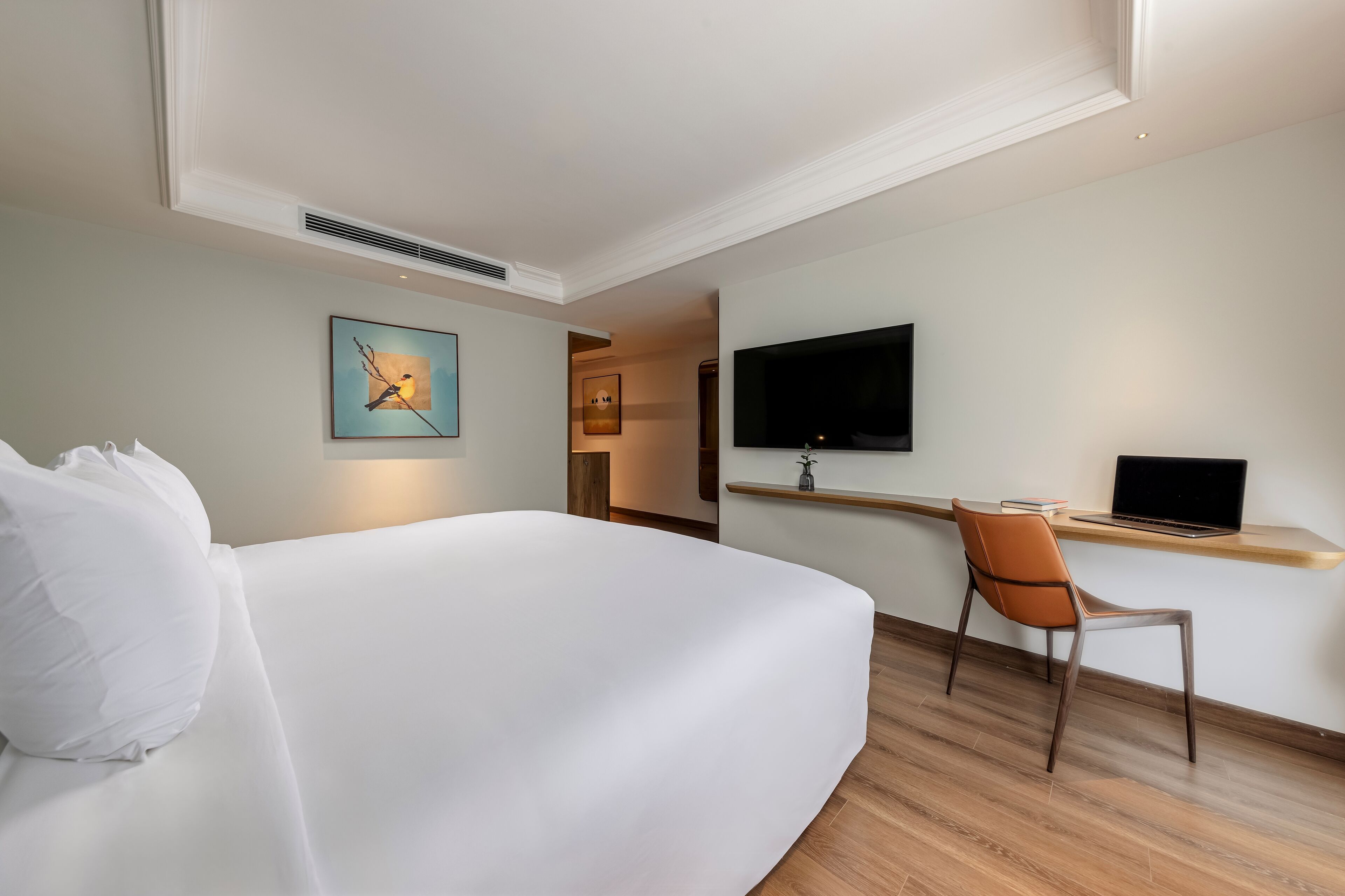 A hotel room with a large bed, a desk with a laptop, a TV mounted on the wall, and artwork on the wall.