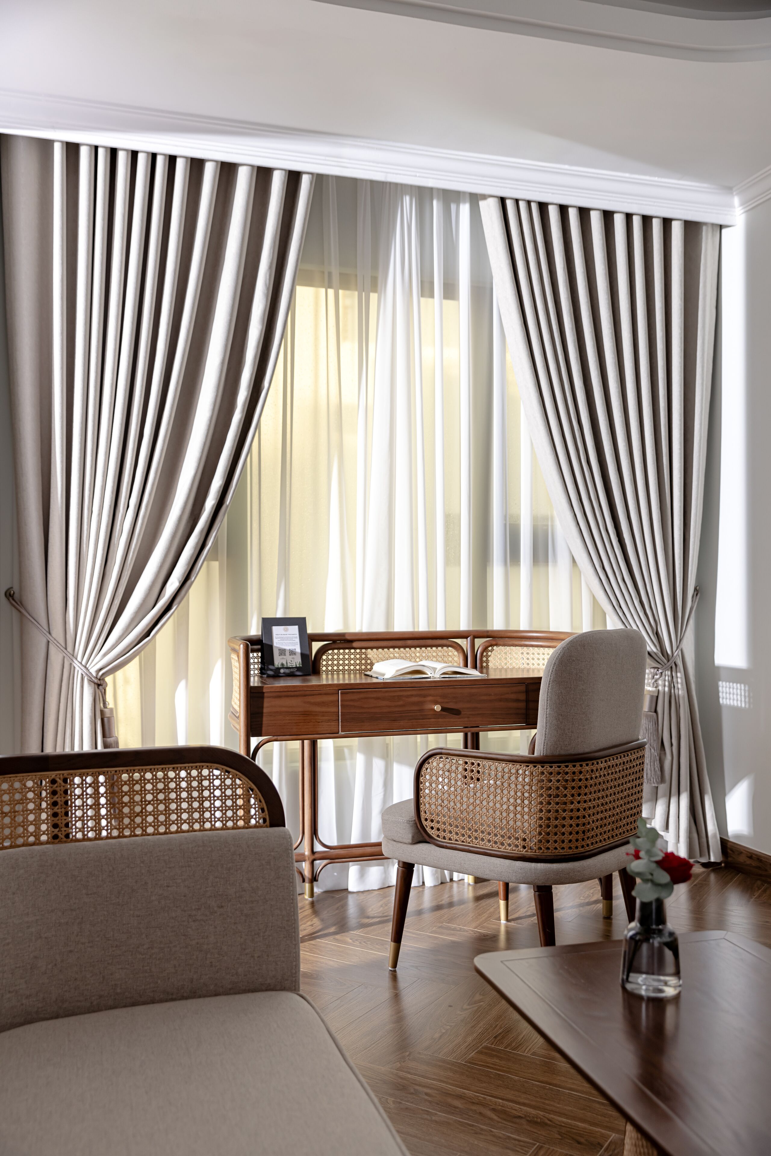 A room with a wooden desk, a wicker chair, a sofa, and a vase with a flower.