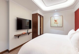 A hotel room with a large bed, a flat-screen TV mounted on the wall, a wooden wardrobe, a framed artwork, and a small shelf with a plant.
