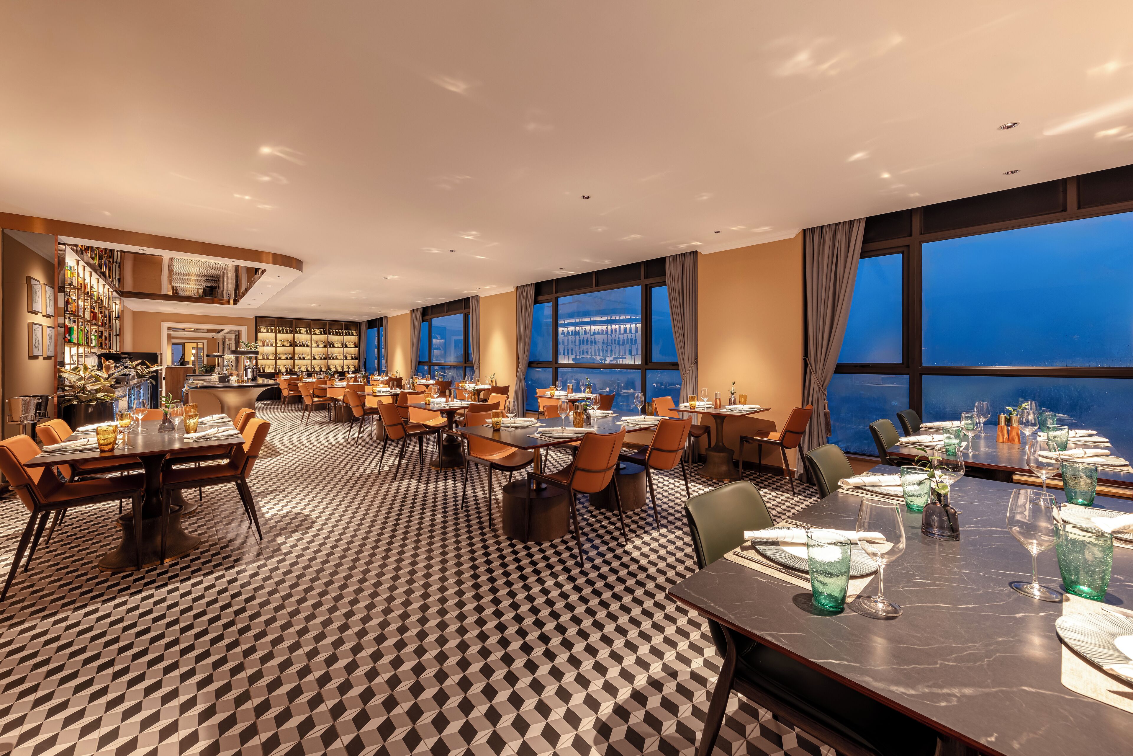 A hotel dining area with multiple tables set for guests, a patterned floor, and large windows offering a view of the cityscape.