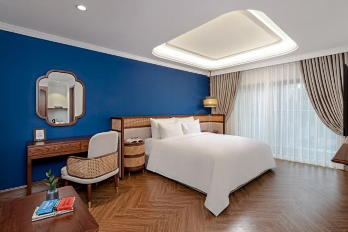 A hotel room with a bed, a desk, a chair, a bedside table, a lamp, a mirror, and a window with curtains.