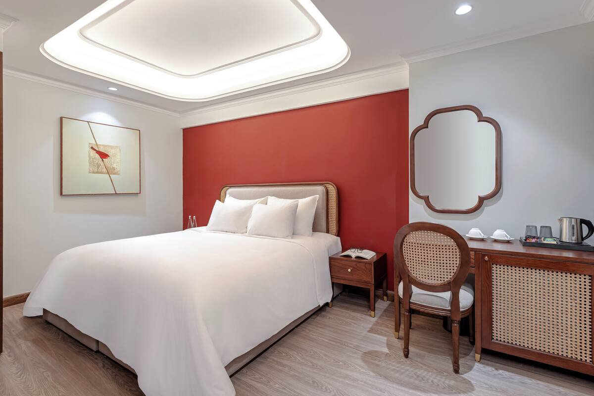 Une chambre d’hôtel avec un grand lit, un bureau avec une chaise, une table de chevet, un miroir et une œuvre d’art encadrée au mur.